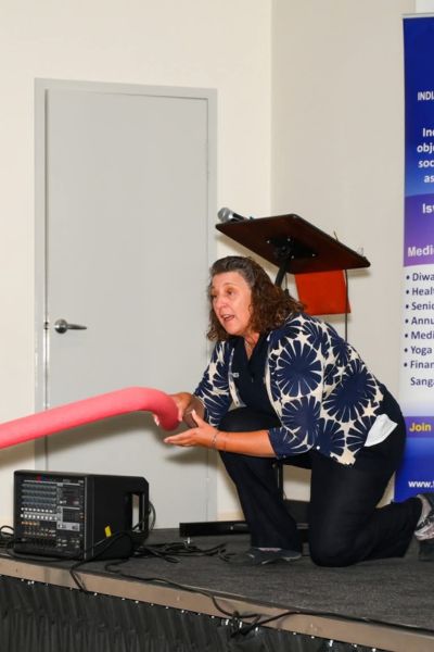 A woman kneels on a stage holding out a pool noodle, demonstrating a reach rescue