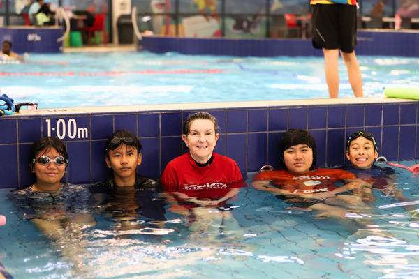 A swim teacher stands between four students as they all stand against the pool wall and smile at the camera