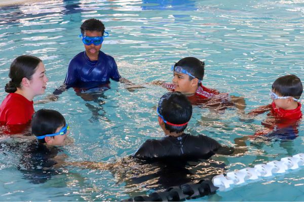 A small group of children wearing goggles gather in a circle in the pool while an instructor speaks to them.