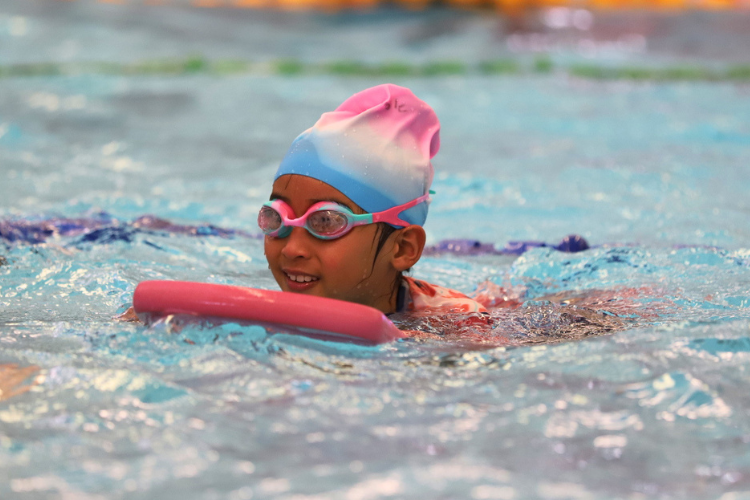 young girl wearing goggles and swim cap using kickboard in pool