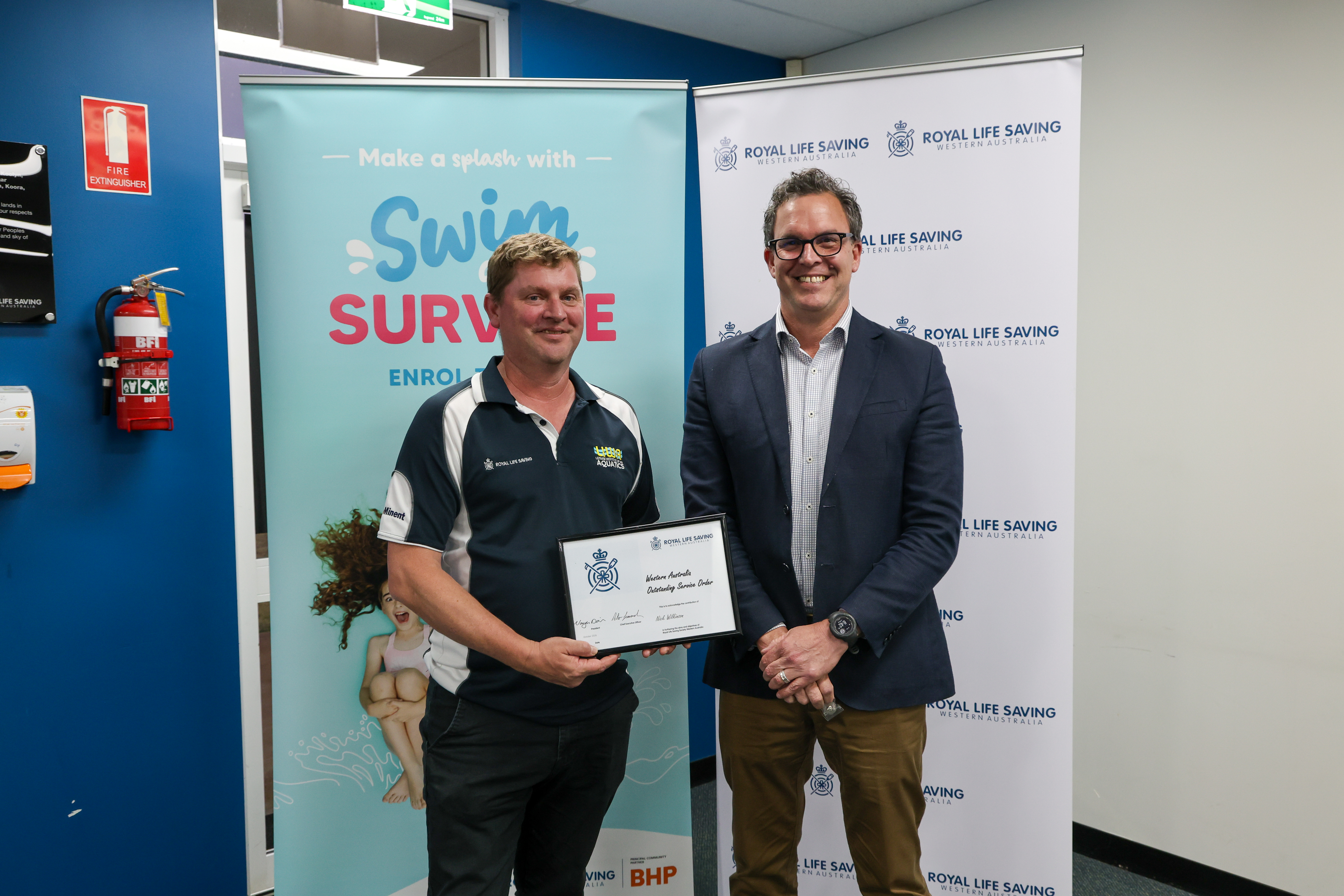 A man holds his Western Australia Outstanding Service Order certificate beside a presenter at Royal Life Saving WA&rsquo;s 2026 AGM.