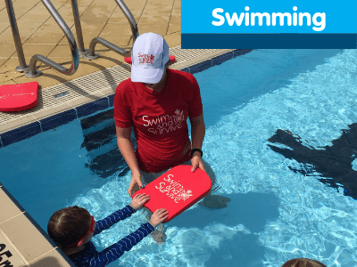 A swim instructor in a pool with a child holding a red Swim and Survive kickboard